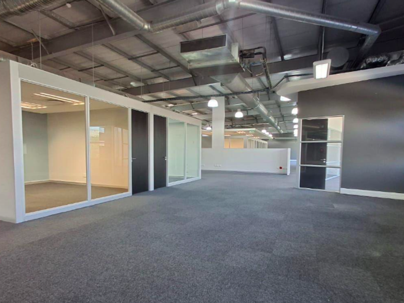 Modern open-plan office space featuring glass-partitioned private offices, exposed industrial-style ceiling ducting, and durable carpet tile flooring.