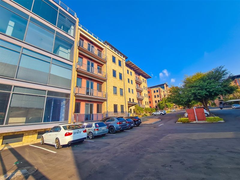 Exterior view of a modern multi-story office building complex featuring glass facades, brickwork, and private balconies, with an adjacent paved parking area.