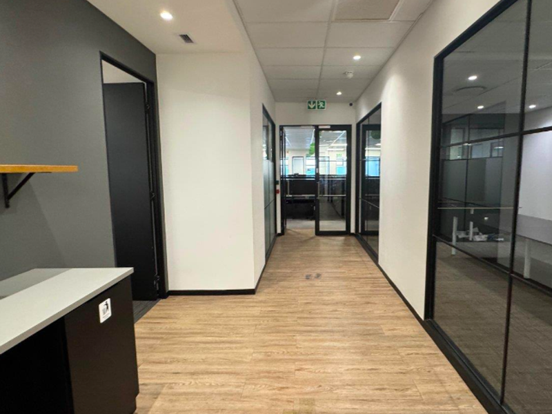 A modern office hallway featuring wood-look laminate flooring, glass partition walls, and recessed ceiling lighting, leading to an open-plan workspace.