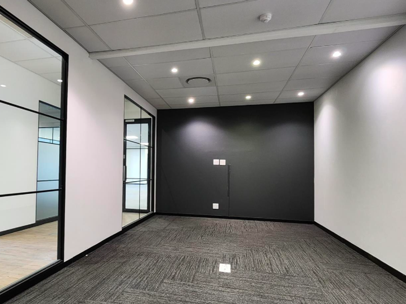 A modern, empty office space featuring glass partitions, a suspended ceiling with recessed lighting, and dark carpet tiles, offering a clean professional environment.