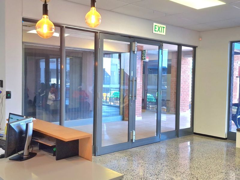 Modern office reception area featuring polished terrazzo flooring, suspended ceiling with recessed lighting, and large aluminium-framed glass doors leading to the exterior.