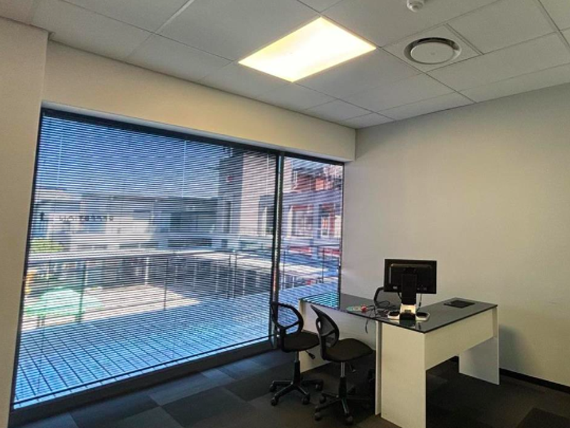 A professional office space featuring a large window with venetian blinds, suspended ceiling with recessed lighting, and carpet tile flooring.