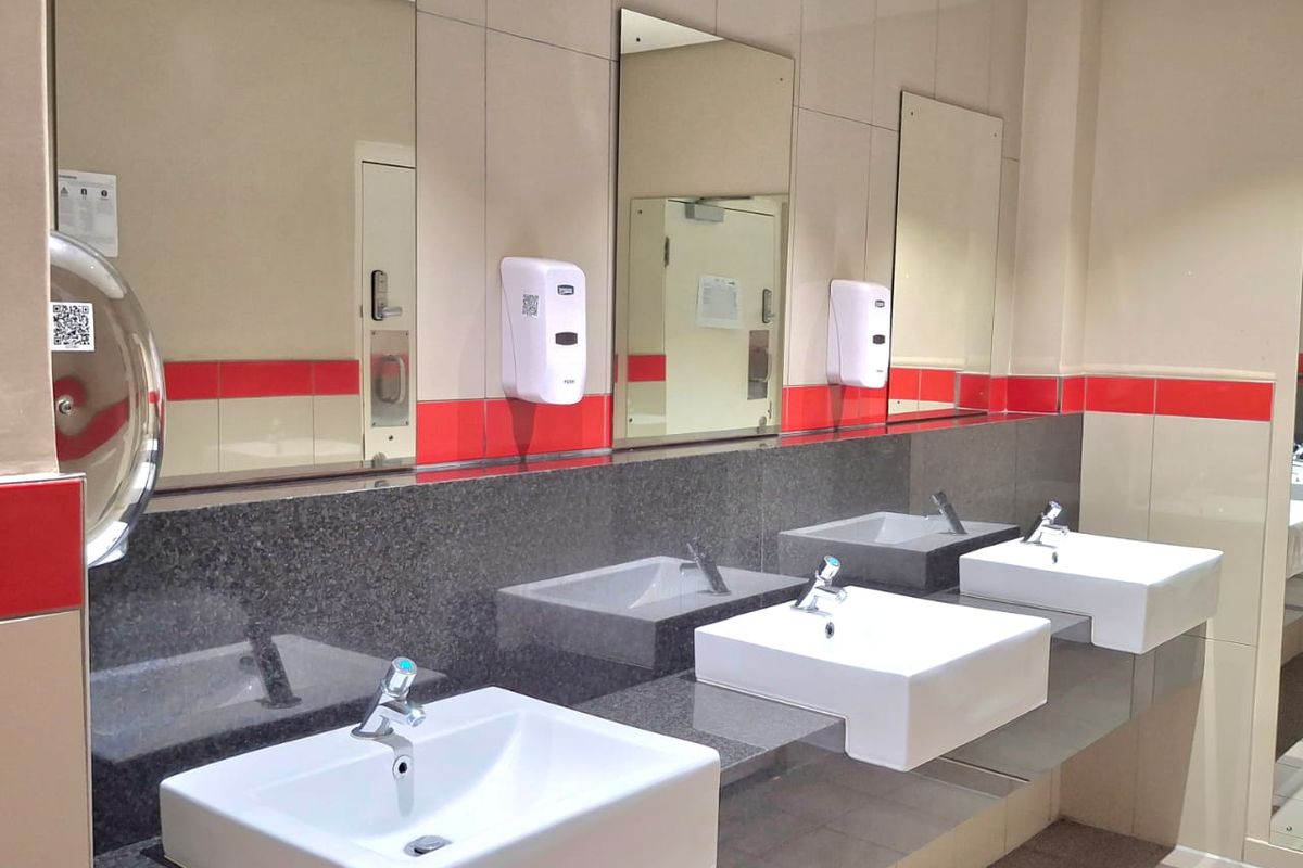 A clean, modern commercial bathroom featuring three wall-mounted ceramic basins, a granite vanity countertop, and wall-mounted mirrors with decorative red tiled accents.