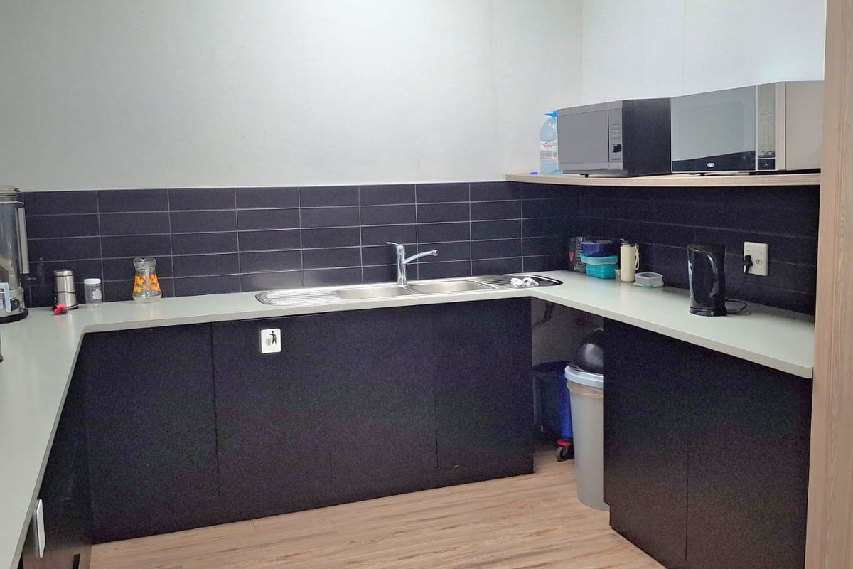 A functional office kitchenette featuring dark cabinetry, a stainless steel sink, tiled backsplash, and light wood-look laminate flooring.