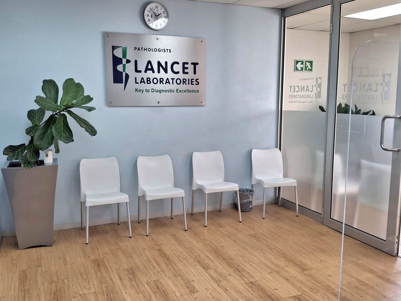 A professional office waiting area featuring light wood-look laminate flooring, a glass partition entrance, and recessed ceiling lighting.