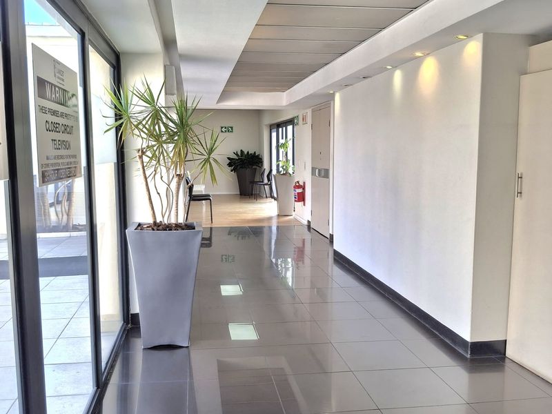 A bright, modern office hallway featuring polished tiled floors, recessed lighting, and large glass panels providing natural light.