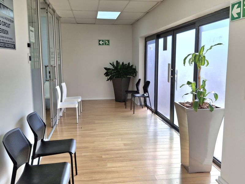 A bright office hallway featuring laminate flooring, suspended ceiling with recessed lighting, and glass partition doors leading to adjacent office spaces.