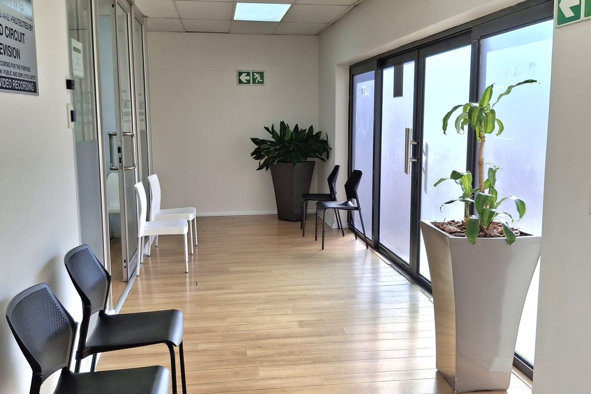 A bright office hallway featuring laminate flooring, suspended ceiling with recessed lighting, and glass partition doors leading to adjacent office spaces.