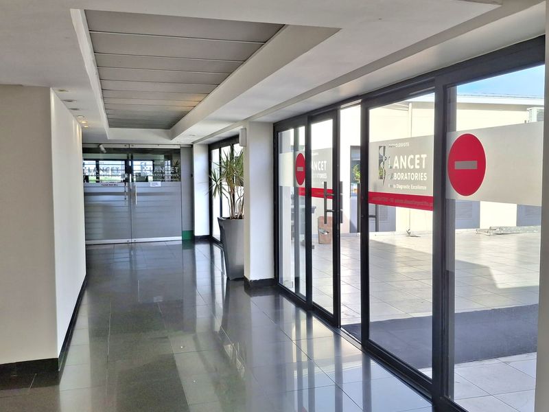A bright, modern office hallway featuring polished tiled flooring, recessed ceiling lighting, and large glass sliding doors leading to an outdoor area.