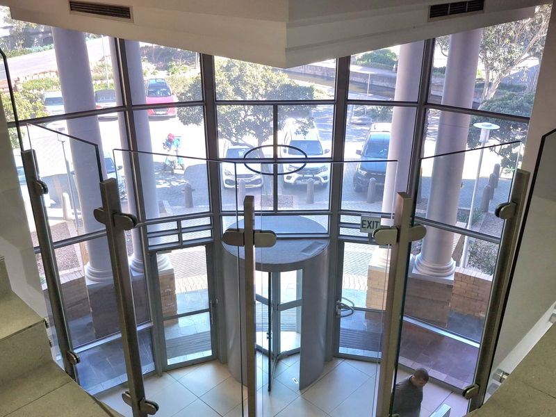 A bright, modern office building entrance featuring a revolving door, floor-to-ceiling glass facade, and a contemporary glass balustrade overlooking the lobby.
