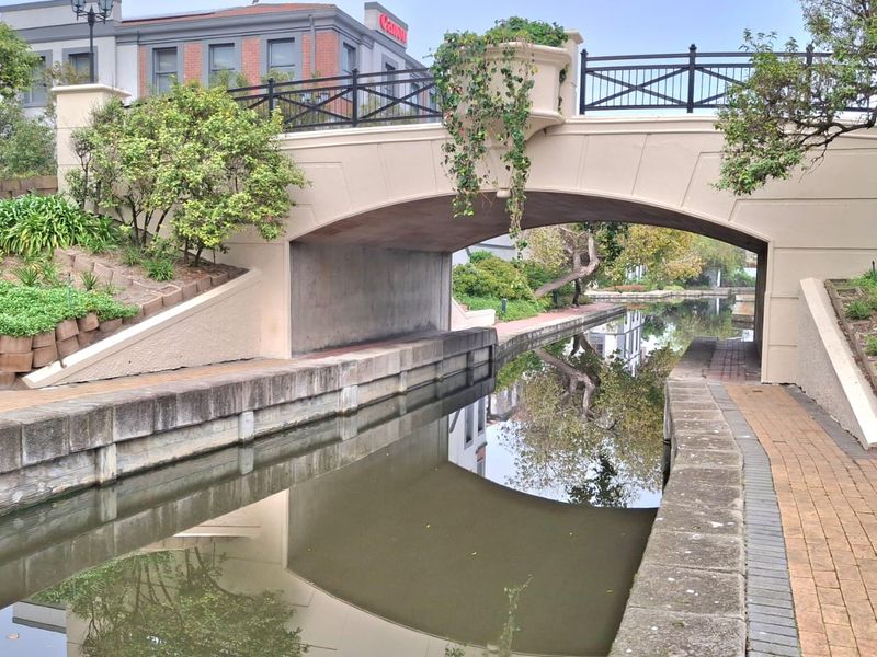 A scenic view of a stone bridge spanning a canal, featuring brick pathways and landscaped retaining walls within the office property grounds.