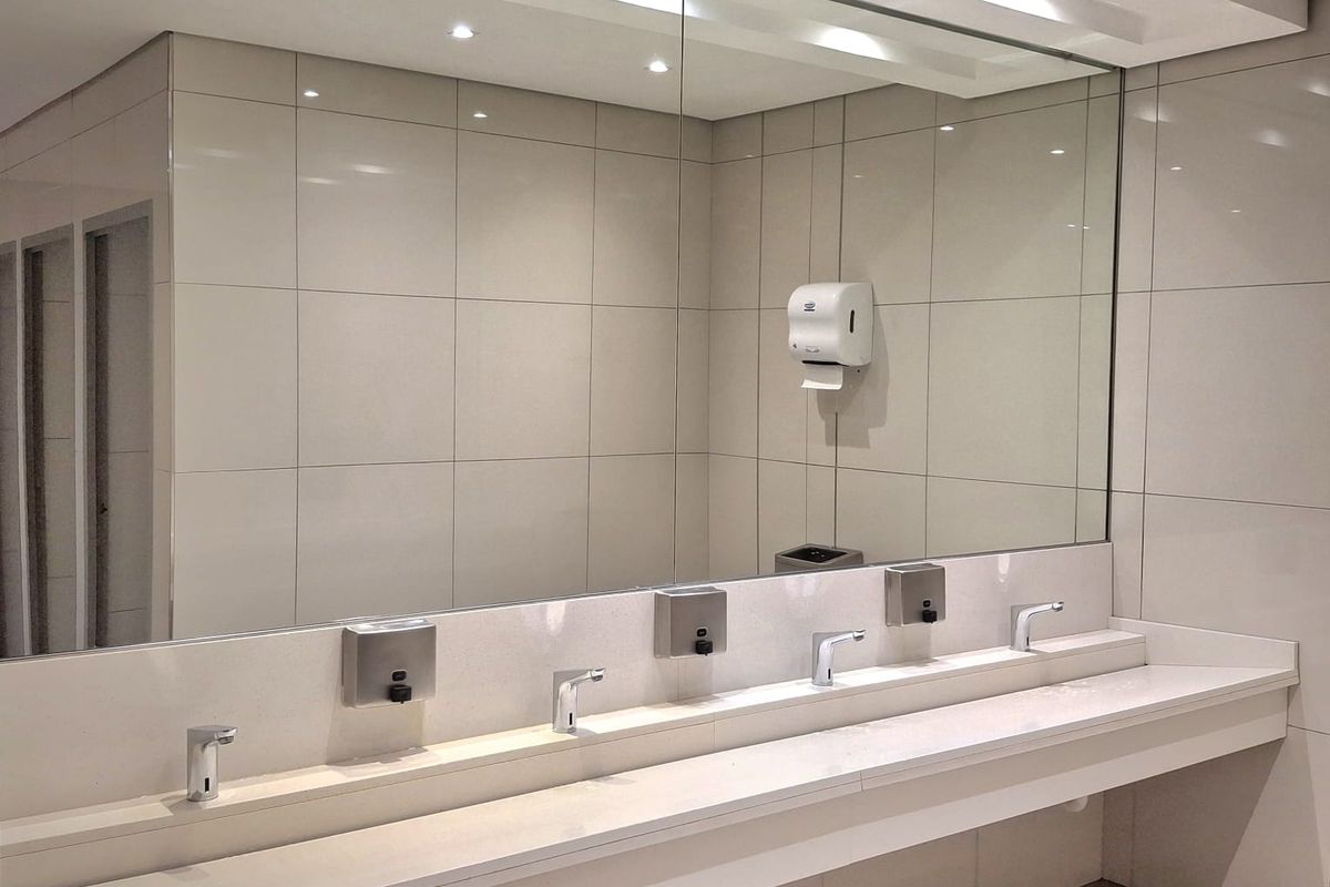 Modern office bathroom featuring a long integrated stone vanity with multiple sensor faucets, large wall-mounted mirror, and clean ceramic tiling.