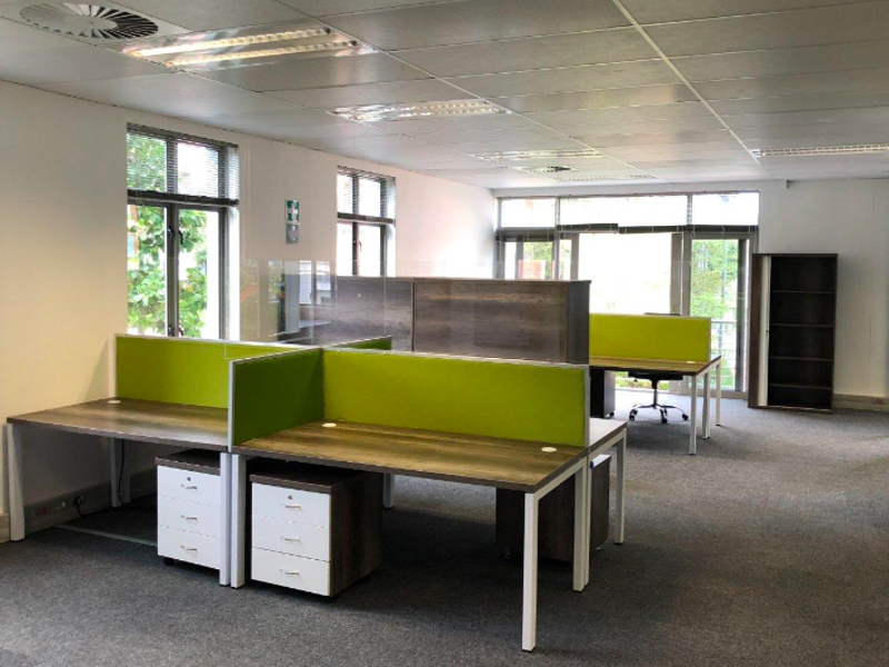 A bright, open-plan office space featuring suspended ceilings, recessed lighting, carpeted floors, and large windows providing ample natural light.