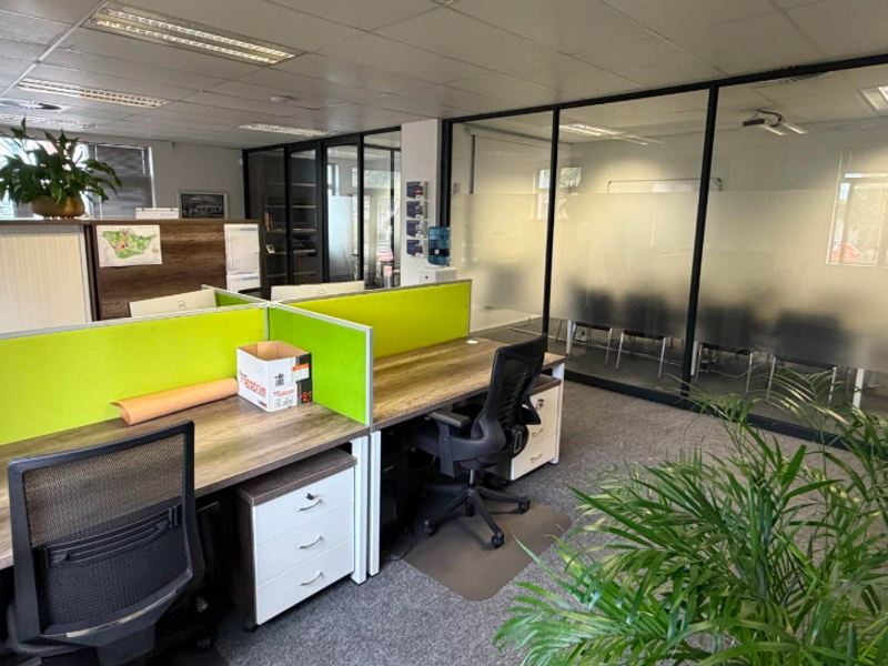 Modern office space featuring glass partition walls, carpeted flooring, and integrated desk workstations with a professional layout.