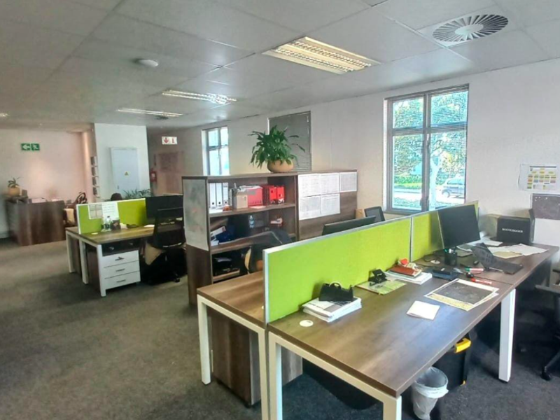 A functional open-plan office space featuring carpeted flooring, suspended ceilings with integrated lighting, and large windows providing natural light.