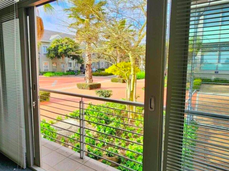 A view from an office interior through a sliding glass door onto a small balcony overlooking a landscaped courtyard.
