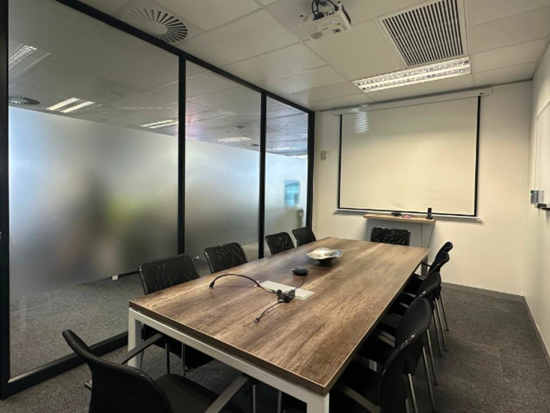 A professional office meeting room featuring a large conference table, glass partition walls, a ceiling-mounted projector, and a retractable projection screen.