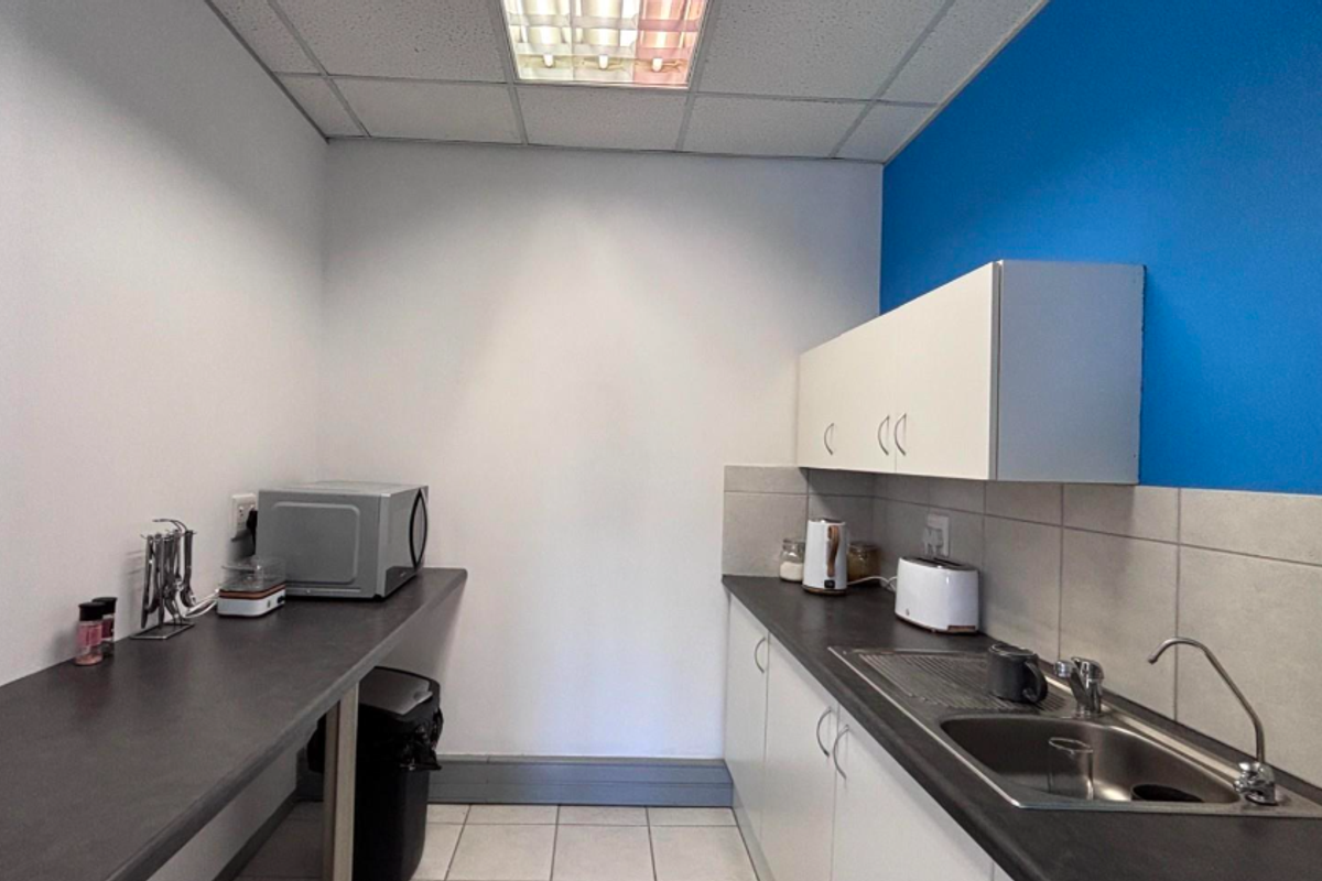 A functional office kitchenette featuring white cabinetry, dark laminate countertops, a stainless steel sink, and a tiled backsplash.