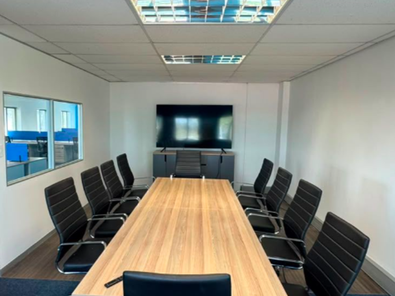 A professional boardroom featuring a large meeting table, suspended ceiling with recessed lighting, and an internal glass partition overlooking an adjacent office space.