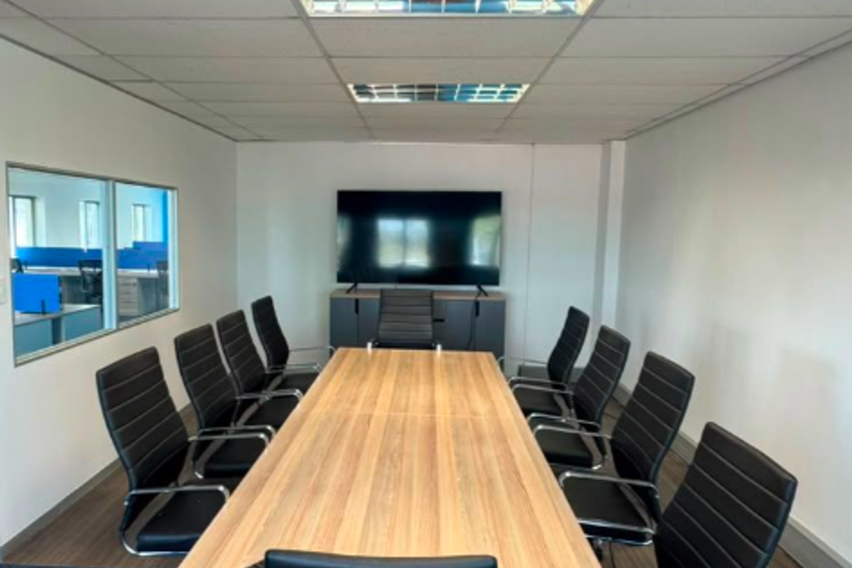 A professional boardroom featuring a large meeting table, suspended ceiling with recessed lighting, and an internal glass partition overlooking an adjacent office space.