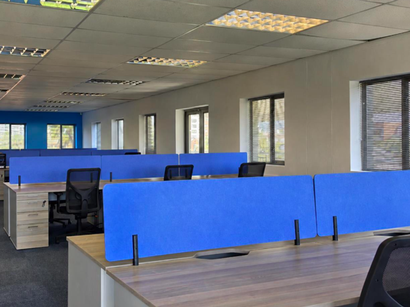 A spacious open-plan office area featuring modular workstations with blue privacy screens, suspended ceilings, and large windows providing natural light.