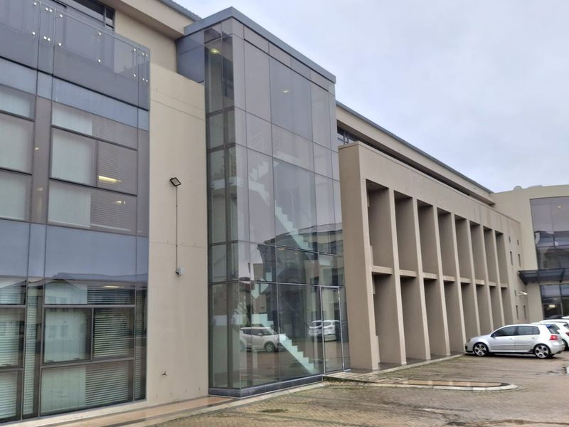 Modern multi-story office building facade featuring a prominent glass-enclosed staircase, concrete structural columns, and an adjacent paved parking area.