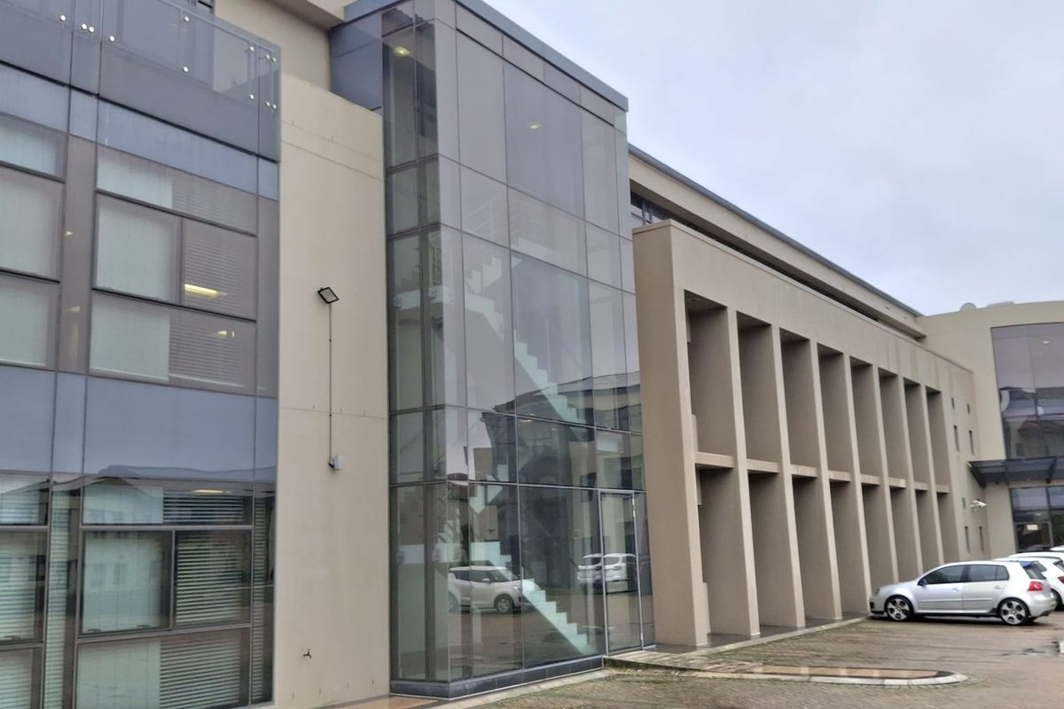 Modern multi-story office building facade featuring a prominent glass-enclosed staircase, concrete structural columns, and an adjacent paved parking area.