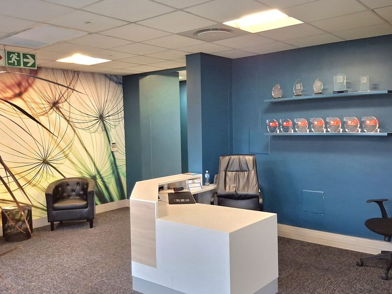 A professional office reception area featuring a modern built-in desk, floating display shelves, carpeted flooring, and a decorative feature wall.