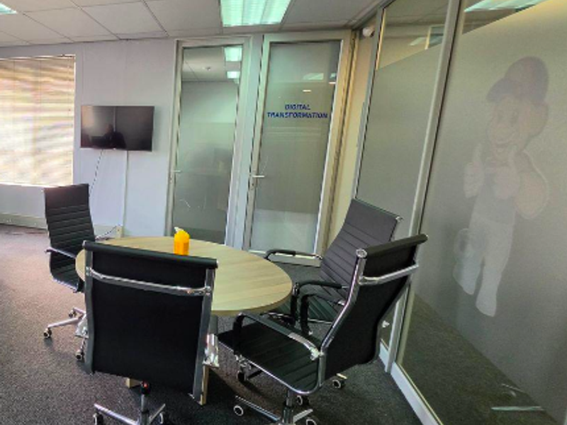 A professional office meeting space featuring glass partition walls, suspended ceiling with recessed lighting, and carpeted flooring.