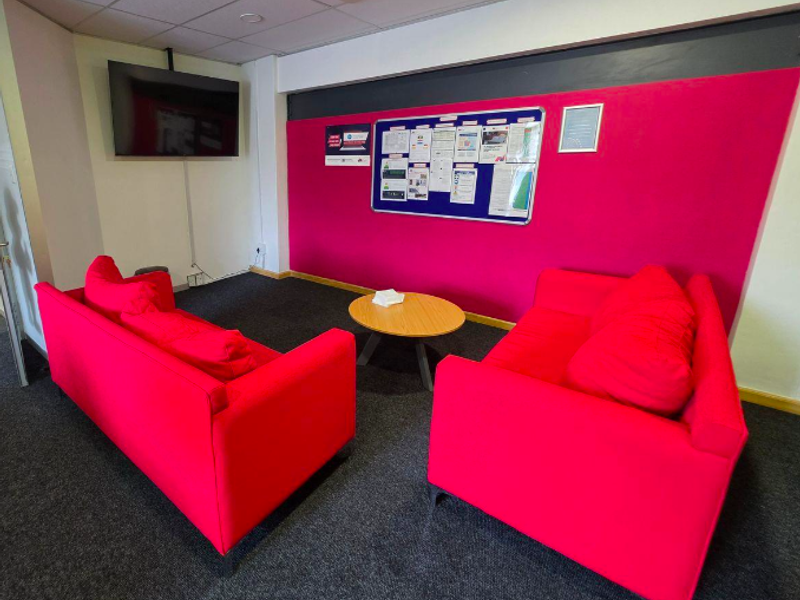 A functional office breakout or waiting area featuring carpeted flooring, a suspended ceiling, and a prominent accent wall.