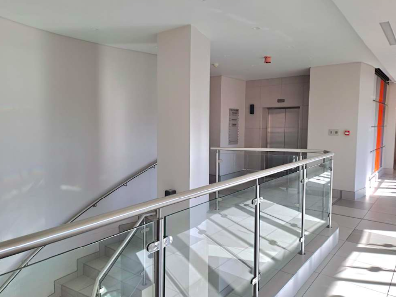 Modern office building hallway featuring a glass balustrade, stainless steel handrail, and elevator access with clean tiled flooring and bright recessed lighting.
