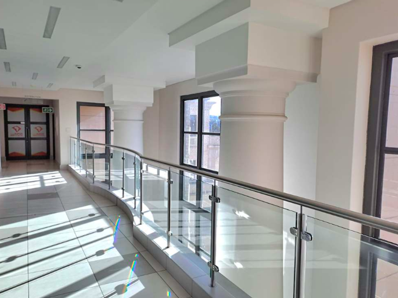 A bright, modern office hallway featuring tiled flooring, a sleek glass balustrade, and decorative columns with recessed ceiling lighting.