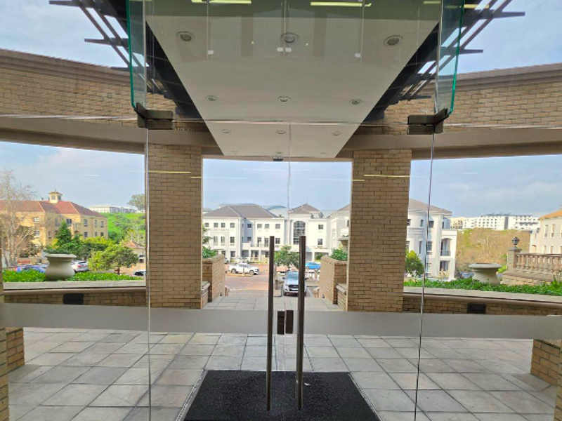 Modern office building entrance featuring glass double doors, brick structural pillars, and stone tile flooring with a view of the surrounding office park.