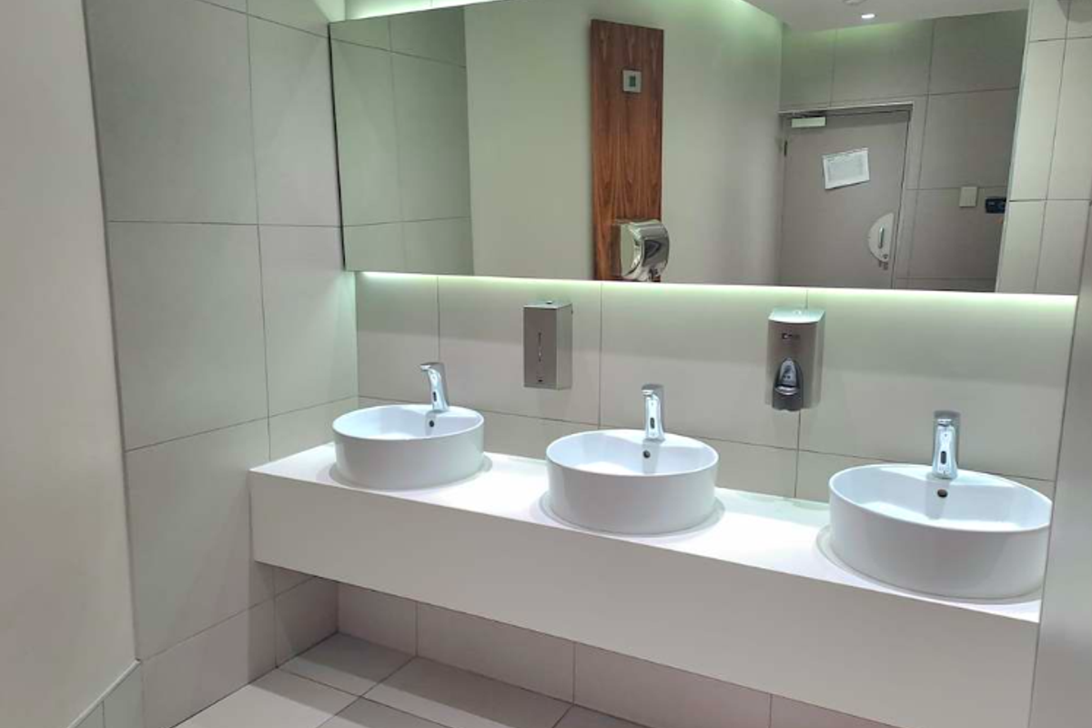 Modern office bathroom featuring a sleek floating vanity with three vessel sinks, wall-mounted faucets, and large illuminated mirror with tiled finishes.