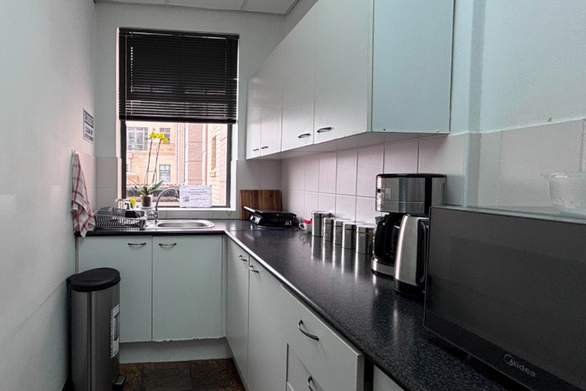 A compact office kitchenette featuring white cabinetry, dark laminate countertops, a stainless steel sink, and a tiled backsplash.