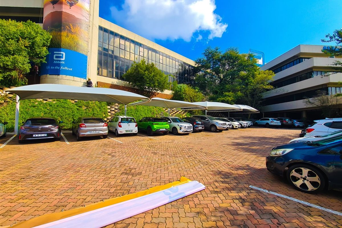 An outdoor parking area featuring shade cloth carports, paved surfaces, and a view of the office building facade with a large advertising banner.