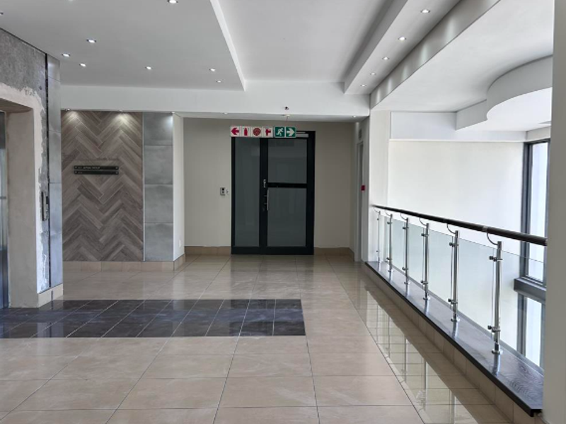 Modern office hallway featuring polished tile flooring, a glass balustrade, recessed lighting, and a decorative herringbone feature wall near the elevator entrance.