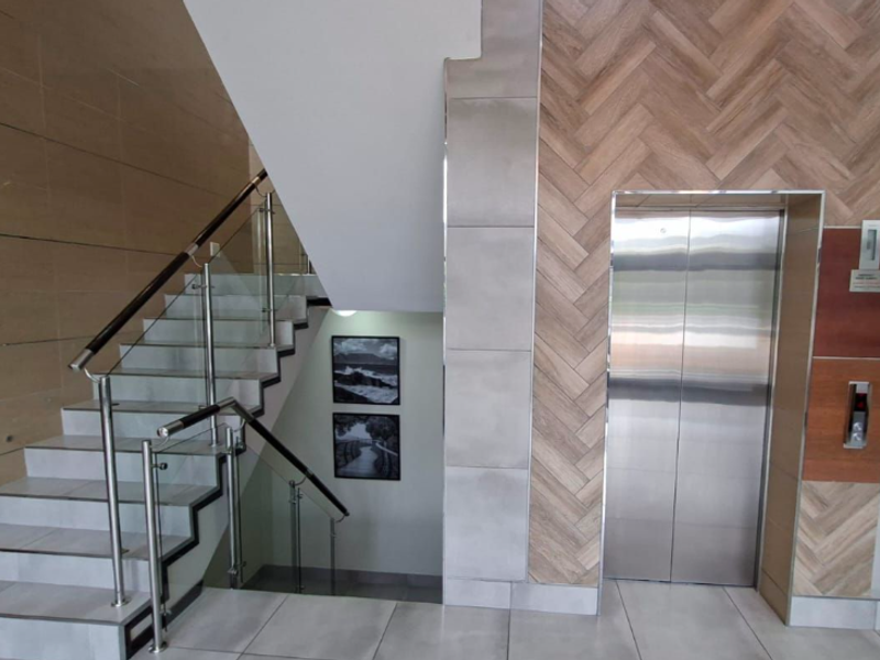 Modern office building lobby featuring a stainless steel elevator, tiled flooring, and a contemporary staircase with glass and steel railings.