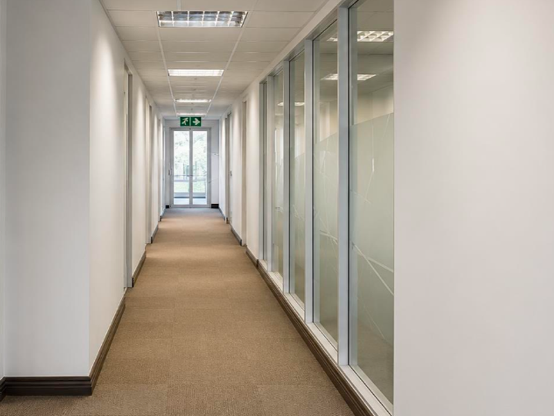 A clean, professional office hallway featuring carpeted flooring, suspended ceilings with recessed lighting, and glass partition walls with frosted privacy detailing.