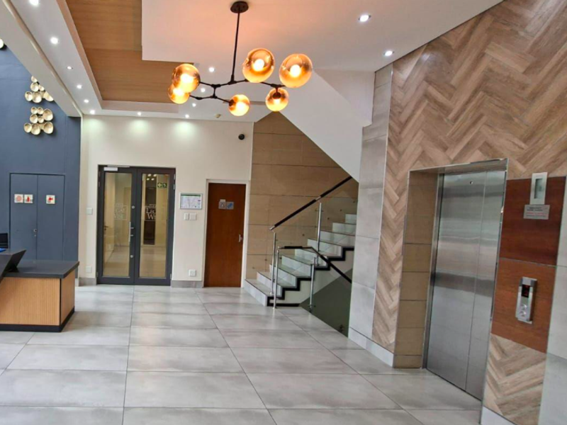Modern office building lobby featuring a reception area, contemporary staircase with glass balustrade, elevator access, and high-end finishes including herringbone wall detailing.