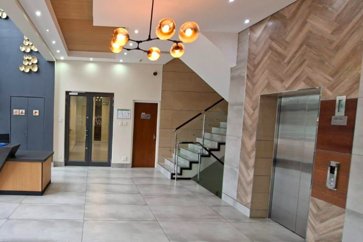 Modern office building lobby featuring a reception area, contemporary staircase with glass balustrade, elevator access, and high-end finishes including herringbone wall detailing.