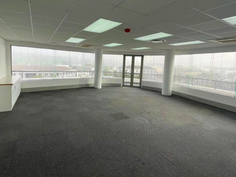 A spacious, empty open-plan office featuring carpet tiles, suspended ceilings, recessed lighting, and expansive windows providing ample natural light and exterior views.