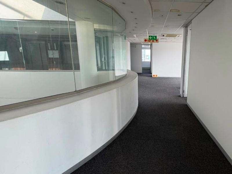A spacious office hallway featuring a curved glass partition, carpeted flooring, and a suspended ceiling with recessed lighting.