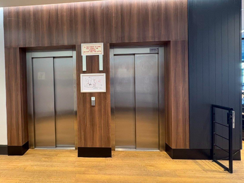 Modern office building elevator lobby featuring dual stainless steel lifts, wood-look wall paneling, and durable laminate flooring.
