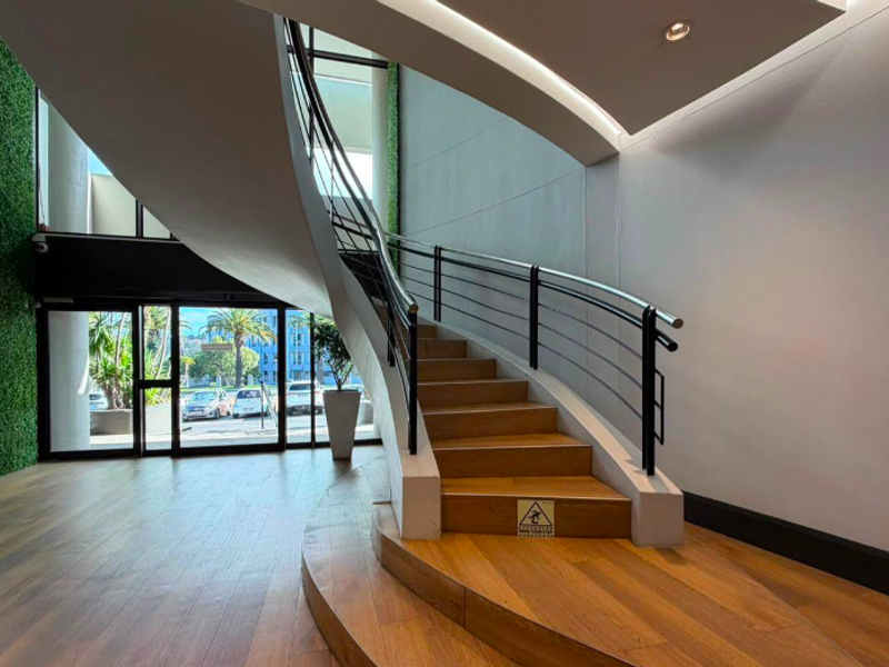 Modern office entrance featuring a striking curved timber staircase, sleek metal handrails, and floor-to-ceiling glass doors leading to the exterior.