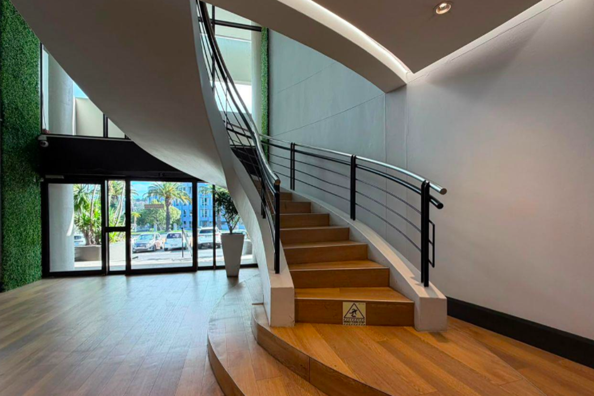 Modern office entrance featuring a striking curved timber staircase, sleek metal handrails, and floor-to-ceiling glass doors leading to the exterior.