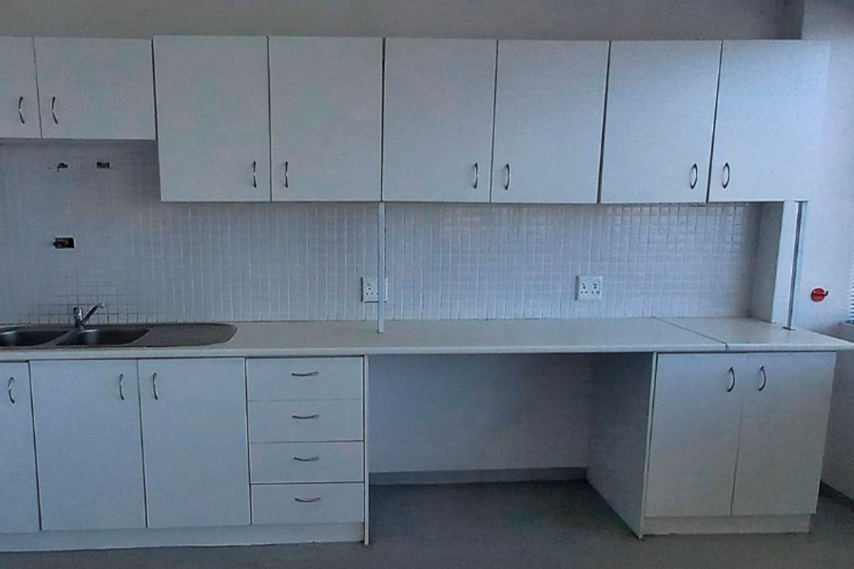 A functional office kitchenette featuring white built-in cabinetry, a stainless steel double sink, tiled backsplash, and ample counter space.