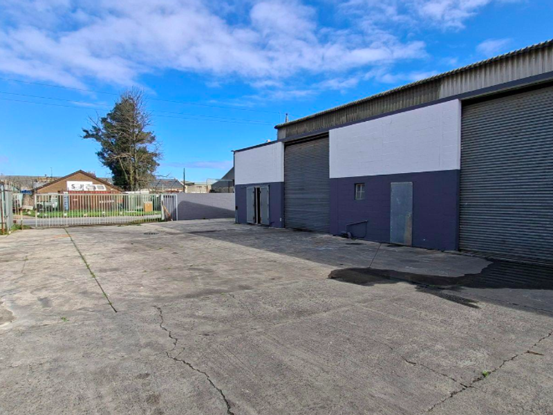 Industrial property exterior featuring a large concrete yard, secure metal fencing, and multiple roller shutter doors for vehicle or goods access.