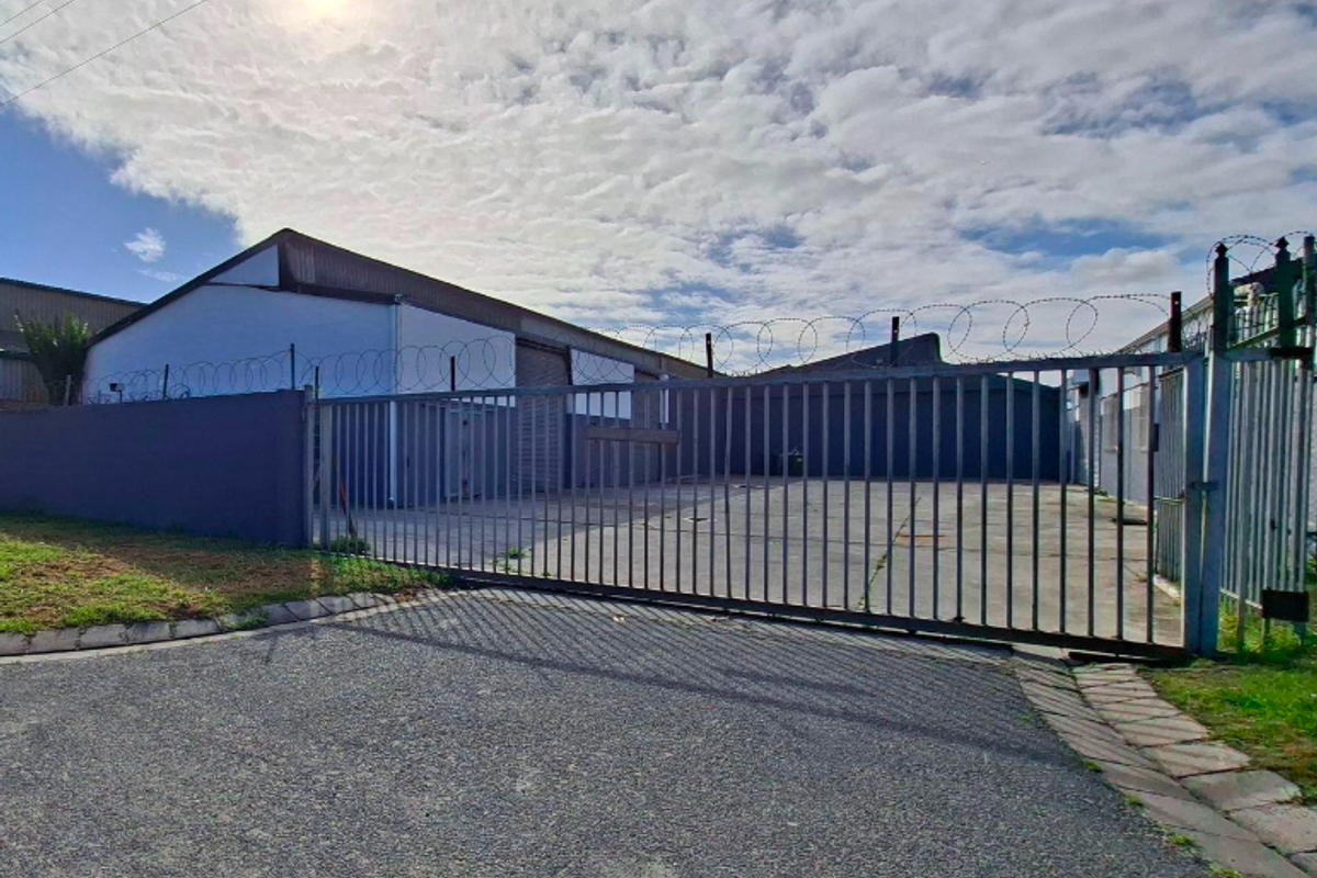 Industrial property exterior featuring a secure gated entrance, concrete driveway, and perimeter wall with razor wire fencing.