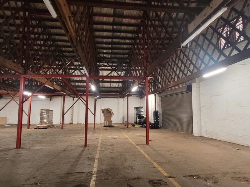 Spacious industrial warehouse featuring high exposed timber truss ceilings, steel support columns, roller shutter door access, and painted brick walls.